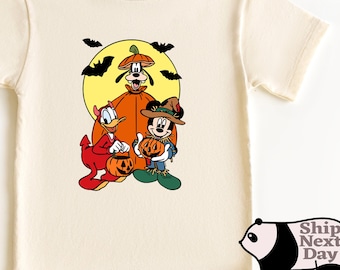 May include: Cream-colored t-shirt featuring a Halloween-themed graphic. The design includes cartoon characters dressed in costumes, including a pumpkin-headed figure, a devil, and a scarecrow. Bats fly in front of a yellow moon.