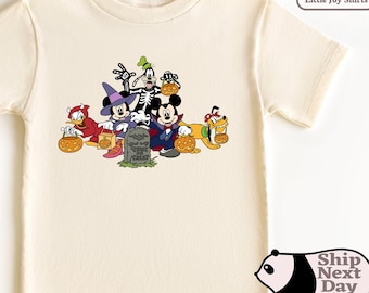 May include: A cream colored short-sleeved t-shirt with a graphic of Mickey Mouse and his friends dressed in Halloween costumes. The graphic includes Mickey Mouse as a vampire, Minnie Mouse as a witch, Goofy as a skeleton, Donald Duck as a devil, and Pluto as a dog wearing a pumpkin.