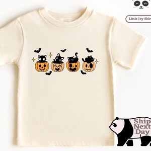 May include: A cream colored short-sleeved t-shirt with a graphic of four black cats sitting in orange jack-o'-lantern pumpkins. The graphic also includes bats and stars.