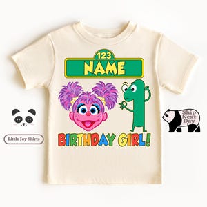 Puede incluir: Camiseta de color crema con un dise&ntilde;o gr&aacute;fico colorido. El dise&ntilde;o incluye un personaje de dibujos animados con pelo morado, el n&uacute;mero 1 y las palabras "BIRTHDAY GIRL!" en colores arco&iacute;ris. La camiseta tambi&eacute;n tiene el texto "NAME".