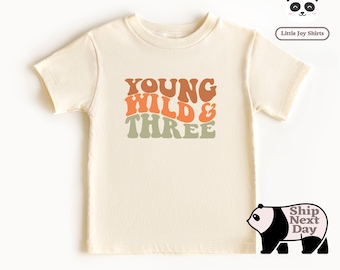 May include: A cream colored short-sleeved t-shirt with the text "YOUNG WILD & THREE" in a retro font with orange, brown, and green colors.