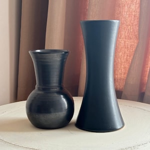 May include: Two dark grey ceramic vases. One is a rounded shape with a narrow neck, and the other is a tall, flared vase. Both vases are on a white table, with a neutral-coloured curtain backdrop.