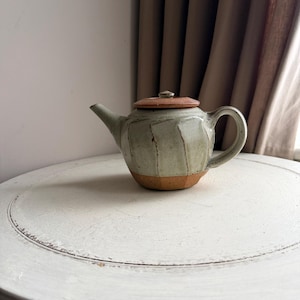 May include: A handcrafted ceramic teapot with a pale green glaze and a brown lid. The teapot features a rounded body with vertical textured lines and a curved handle. The base is a warm brown colour. A small, round knob sits atop the lid.