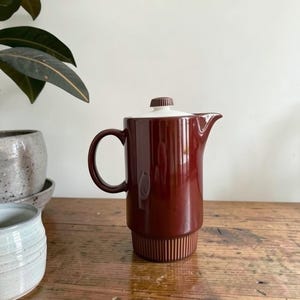 May include: A cylindrical ceramic coffee pot featuring a deep brown glossy finish on the upper body and a darker brown vertically ribbed base. It includes a white lid with a small brown knob handle and a curved handle on the side.