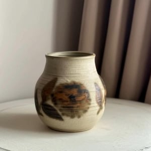 May include: A beige ceramic vase with a rounded body and a slightly flared rim. The vase is decorated with brown and black floral patterns. The vase is sitting on a white surface.