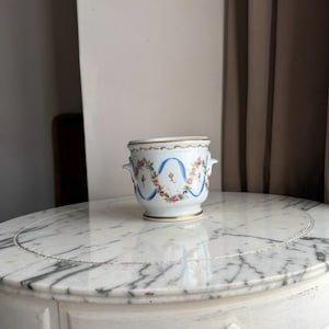 May include: A white porcelain cachepot featuring a hand-painted floral motif with pink and orange flowers, green leaves, and blue ribbon swags. The vessel has two small side handles and is accented with gold trim along the rim and the base.