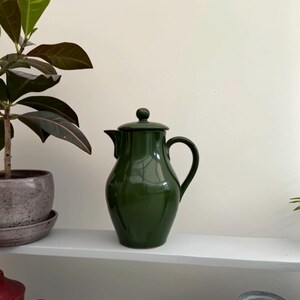 May include: A dark green ceramic coffee pot with a lid and a curved handle. The pot is displayed on a white shelf, with a potted plant to the left. The pot has a classic, elegant design.