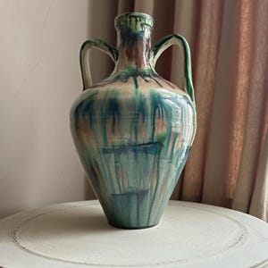 May include: A tall ceramic vase with two handles, in teal, green, and brown. The vase has a bulbous body and a narrow neck. The glaze has a dripped effect. The vase is displayed on a white, round table. The table is approximately 76 cm in diameter.