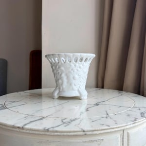 May include: A white ceramic planter featuring a flared, openwork lattice rim and a body decorated with embossed floral patterns. The vessel is supported by three small, curved feet, providing a stable base for this decorative container.