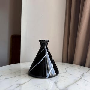 May include: A conical glass vase featuring a glossy black finish with thin, white diagonal swirling lines that wrap around the body, leading up to a narrow, circular opening at the top of the vessel, creating a distinct contrast in colour.