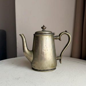 May include: A vintage silver-coloured teapot with a curved spout and handle. The teapot has a lid with a small decorative finial. The pot is sitting on a white surface. The overall design is classic and elegant.