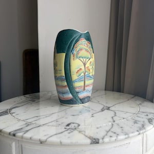 May include: A cylindrical ceramic vase featuring a dark green vertical band and a yellow panel decorated with a painted landscape of trees and rolling hills in shades of orange, blue, and green. The vase has a smooth finish and a curved top edge.