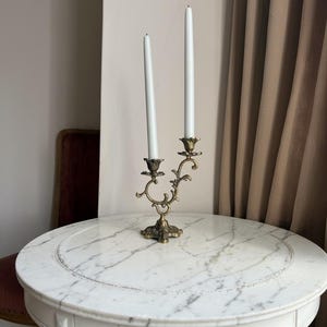 May include: A dual-arm brass candelabra featuring an ornate, curved metal frame with floral-inspired bobeches at each candle cup. Two slender, white taper candles are inserted into the holders, standing at slightly different heights.