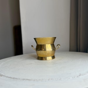 May include: A small, polished brass vessel featuring a wide, flared rim, a rounded body, and two small, curved handles on opposite sides. The metal has a warm, golden tone with a smooth, reflective surface and a sturdy, circular base.