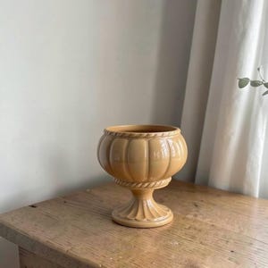 May include: A light tan ceramic pedestal bowl featuring a rounded, ribbed body with a rope-textured rim and a flared, scalloped base. The bowl has a smooth, glossy finish and a wide, open top, standing on a short, sturdy pedestal stem.