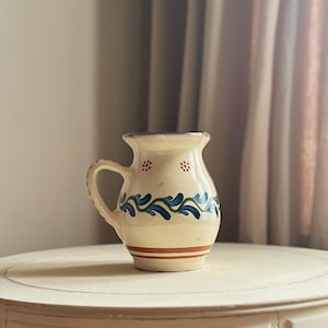 May include: Cream-coloured ceramic jug with a handle, decorated with blue and green floral patterns and a red stripe. The jug is displayed on a white, round table, showcasing its rustic charm and handcrafted design. The jug is a decorative item.