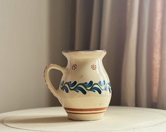 May include: Cream-colored ceramic pitcher with a handle, adorned with blue and green floral patterns and a red stripe. The pitcher is displayed on a white, round table, showcasing its rustic charm and handcrafted design. The pitcher is a decorative item.