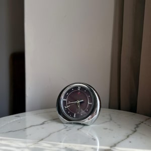May include: A round analogue alarm clock with a polished chrome finish and a dark brown face. The clock features silver-toned hour markers, white numerals at the 12 and 6 positions, and thin silver hands. A small red indicator is visible near the top.