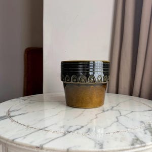 May include: A cylindrical ceramic planter featuring a two-tone glaze. The upper section is a deep, dark green with a repeating embossed arch pattern, while the lower portion transitions into a warm, mottled golden-brown finish with a smooth texture.