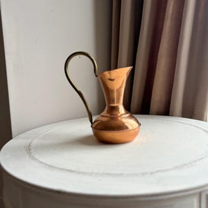 May include: A small, copper-coloured jug with a brass handle. The jug has a rounded base and a flared top. It sits on a white, round table. The handle curves upwards and then back down to meet the jug's body.