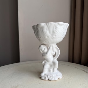May include: A white ceramic decorative bowl with a cherub figurine base. The bowl has a scalloped rim and a detailed leaf pattern. The cherub is holding the bowl aloft. The piece is on a white table.