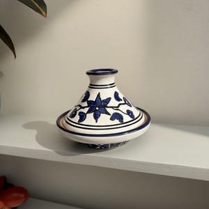May include: A white and blue ceramic tagine pot with a conical lid. The pot features a floral design in blue, with black and brown accents. The tagine sits on a white shelf.