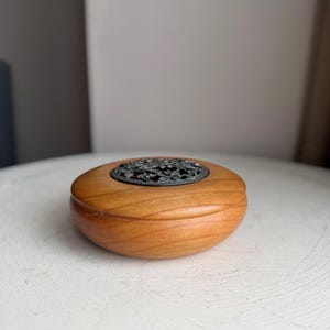 May include: A small, oval wooden box with a decorative metal top. The box is made of light brown wood and has a dark, ornate metal design on the lid. The box is sitting on a white surface.