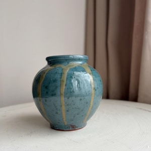 May include: A small, round ceramic vase in shades of teal and beige. The vase has a bulbous shape with a narrow opening and vertical beige drips. The vase is sitting on a white surface.
