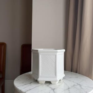 May include: A white ceramic planter with a hexagonal shape and a raised diagonal ribbed texture on each side. The planter features a wide rim, a smooth finish, and stands on small, integrated feet at the base, providing a stable foundation.