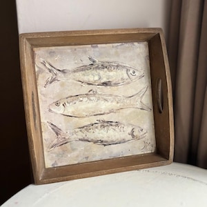May include: A rectangular wooden serving tray with handles. The tray displays a painting of three fish in white and grey tones on a textured surface. The frame of the tray is a warm brown colour, suitable for serving food or drinks.