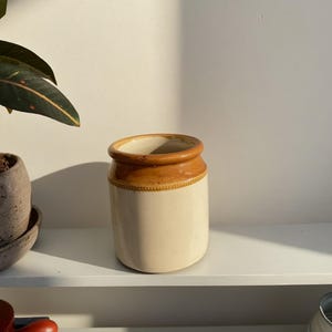 May include: A cream-coloured ceramic jar with a brown rim and accents. The jar is cylindrical with a slightly rounded base. It sits on a white shelf, with a plant and other objects in the background.