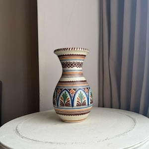 May include: A decorative ceramic vase with a cream base and a flared top. The vase features intricate patterns in brown, blue, green, and orange. The vase is approximately 20 cm tall and sits on a white table.