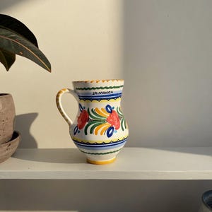 May include: A white ceramic jug with a handle, decorated with red, yellow, and blue floral patterns. The jug has the text "LA MANGA" in blue. The rim and base are trimmed with yellow and blue.