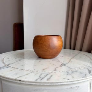 May include: A small, rounded wooden bowl with a smooth, polished finish and visible natural grain patterns in a warm, medium-brown tone. The bowl features a wide, open top and a curved, spherical body that tapers slightly towards the base.