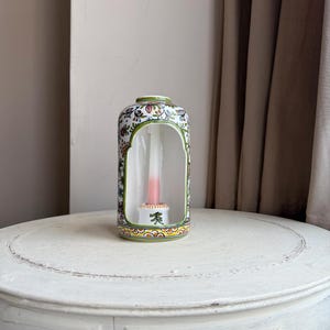 May include: A decorative ceramic candle holder with a floral pattern and a green rim. The candle holder has a rectangular shape with an arched opening and contains a pink and white candle. The candle holder is on a white table.
