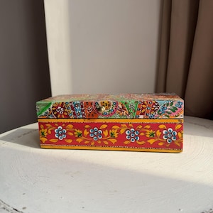 May include: A decorative wooden box with a hinged lid, adorned with a colourful floral pattern. The box is predominantly red, with accents of yellow, blue, and green. A small brass knob secures the lid. The box sits on a white, distressed table, creating a vintage feel.