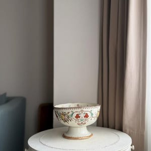 May include: A cream-coloured ceramic pedestal bowl featuring a scalloped rim and a decorative floral pattern in shades of red, yellow, and green. The bowl has a wide, stable base with a gold-toned trim along the bottom edge and the upper rim.