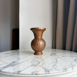 May include: A small, metallic copper-coloured vase with a rounded, bulbous body and a flared rim. The surface of the vase features a distinct hammered texture, creating a series of small, uniform indentations across the entire exterior.