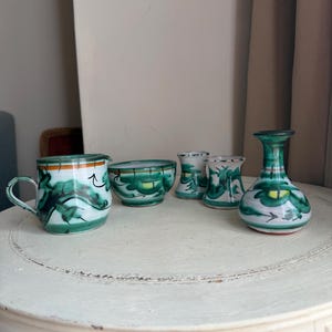 May include: A set of hand-painted ceramic items, including a mug, bowl, and vases. The pieces have a white base with green and orange floral patterns. The objects are displayed on a white, round table.