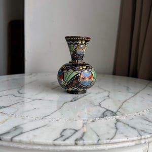 May include: A small, round ceramic vase with a flared neck, featuring a dark black base colour decorated with intricate, colourful patterns in shades of light green, blue, orange, and yellow, including stylised circular motifs around the body.