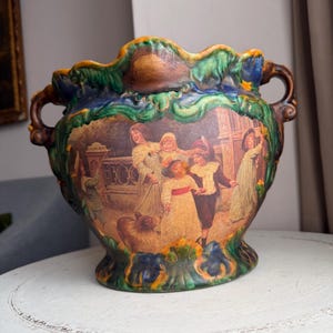 May include: A decorative ceramic planter with a scalloped rim and two handles. The planter features a painted scene of people in period clothing. The rim is green and brown, with blue and yellow accents. The planter is on a white surface.