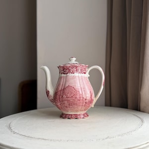 May include: A decorative red and white ceramic teapot featuring a detailed landscape scene. The teapot has a curved spout, a handle, and a lid. It is displayed on a white, round table.