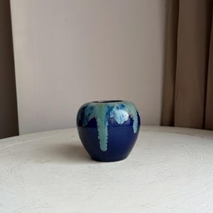 May include: A small, round, navy blue ceramic vase with a glossy finish. The top edge and upper portion of the vase feature a turquoise and white dripped glaze design. The vase is sitting on a white surface.