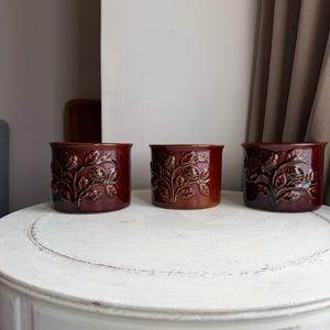 May include: Three cylindrical ceramic planters in a deep, glossy brown glaze. Each planter features a raised, embossed relief pattern depicting rose flowers and leaves wrapping around the exterior surface of the vessel.