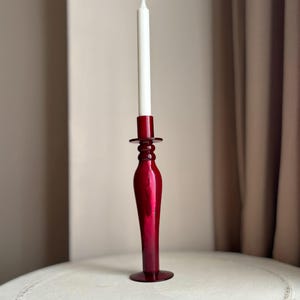 May include: A deep red glass candlestick with a white candle. The candlestick has a flared base, a slender stem, and a circular top. The white candle is cylindrical. The candlestick is on a white surface. The overall height is approximately 25 cm.