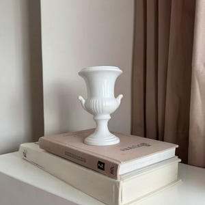 May include: A small, white ceramic urn-shaped vase with a flared rim, a ribbed body, and two small side handles. The vase features a pedestal base and a smooth, glossy finish, standing upright as a decorative object.
