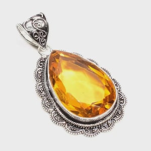 May include: A silver pendant with an ornate bail and a large, teardrop-shaped, golden-yellow gemstone. The gemstone is set in a detailed, silver filigree frame with a scalloped edge. The pendant is displayed against a white background.