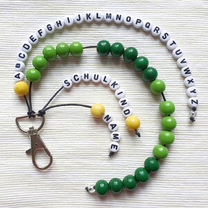 以下が含まれることがあります： アルファベットと「SCHUL KIND NAME」の文字が書かれた白い文字ビーズが付いたビーズのランヤード。 ランヤードには、緑と黄色の木製ビーズと、銀色の留め金も付いています。