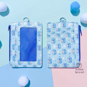 May include: A light blue cardholder with a zipper closure and a silver ring. The cardholder features a repeating pattern of a cartoon character and snowflakes. The background is a mix of light blue and pink.