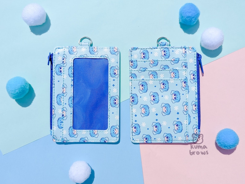 May include: A light blue cardholder with a zipper closure and a silver ring. The cardholder features a repeating pattern of a cartoon character and snowflakes. The background is a mix of light blue and pink.
